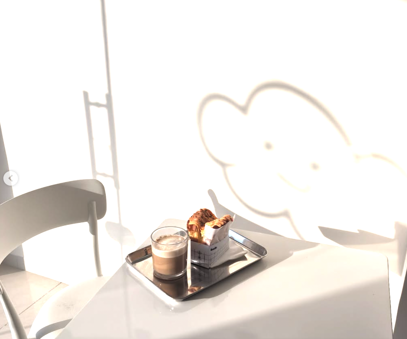 Sunlit café table at Bado with a latte and a Korean sandwich on a silver tray, cloud mascot shadow on the wall